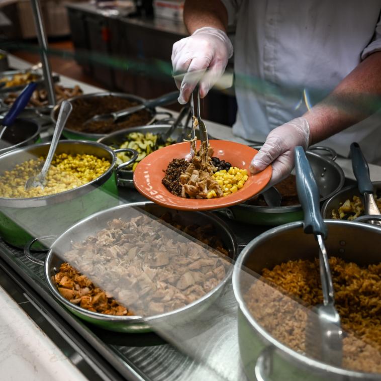 The gloved hands of a cafeteria worker are shown spooning portions of corn, beans, quinoa and shredded meat onto a plate from a buffet 