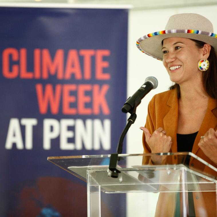 a speaker holds her hands up mid-speech at at a podium. Behind her is a sign that reads "Climate Week at Penn"