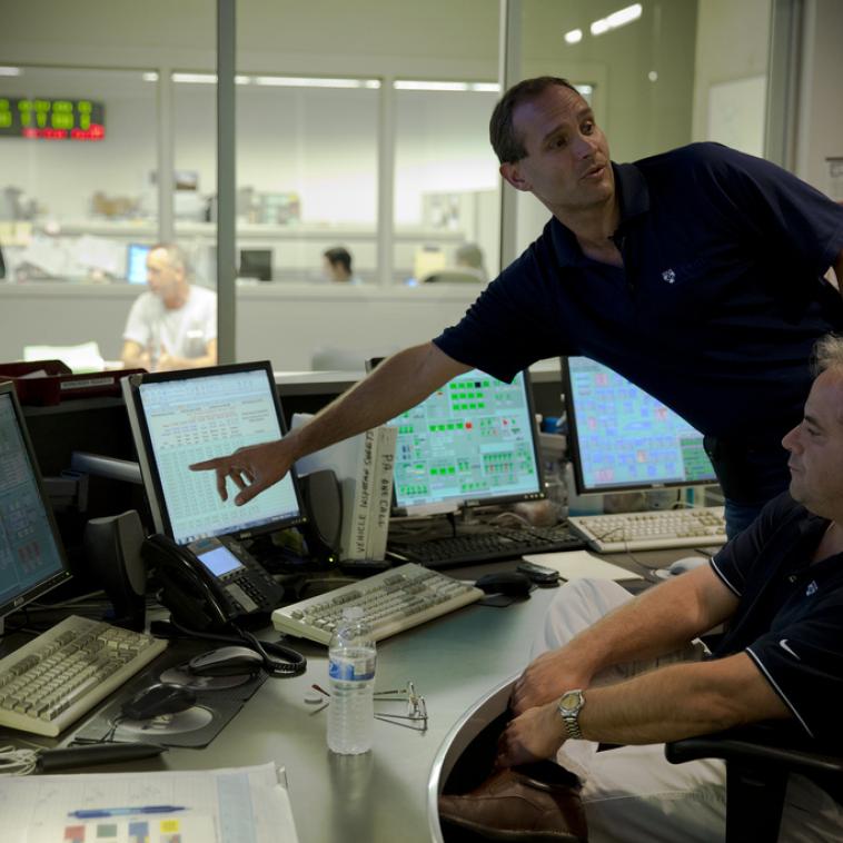 Two people are monitoring computers in control room. One is pointing at a screen and speaking to someone outside of the picture frame.
