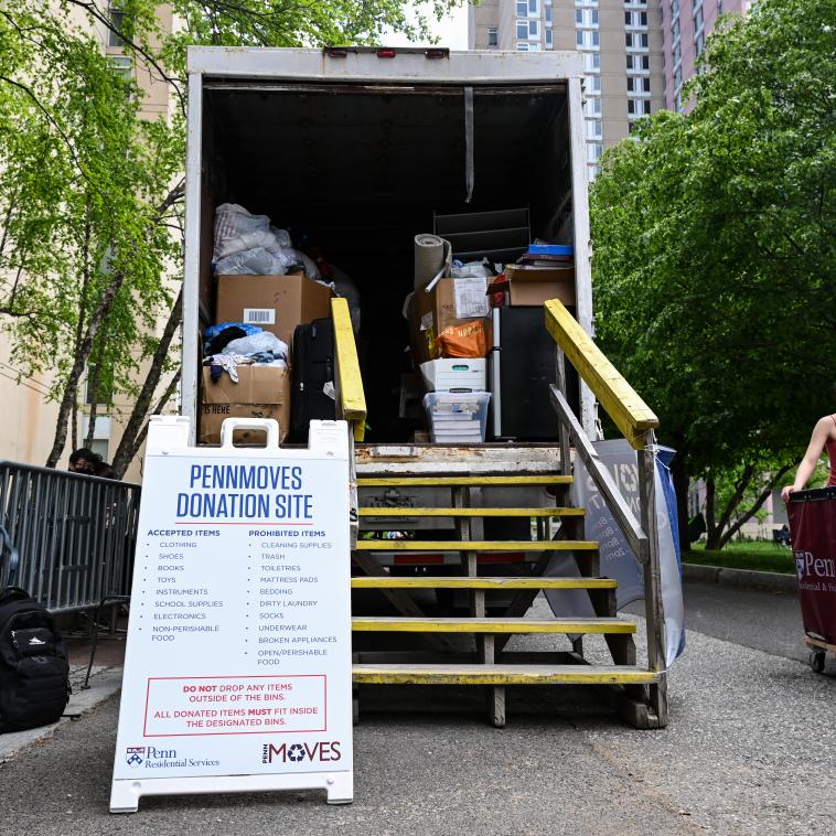 A truck with the back opened is seen parked with steps leading up to the inside. The truck is filled with boxes of assorted items. A sign stands by the staircase leading up to the truck which reads "PENNMOVES DONATION SITE". The sign lists accepted and prohibited items.