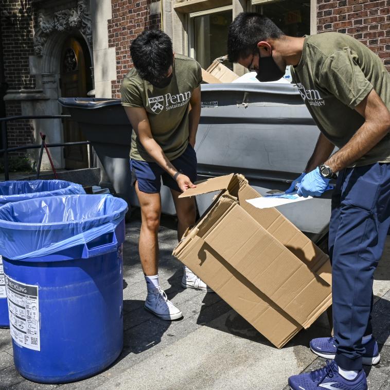 Two students wearing T-Shirts that read "Penn Sustainability" and masks are standing over a cardboard box next to a pair of recycling bins. Behind them is a full recycling dumpster with broken-down cardboard.