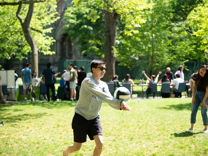 Students laugh while playing vollyball in a field. Behind them tables are set up along the sidewalk for an event and more students are gathered there. 