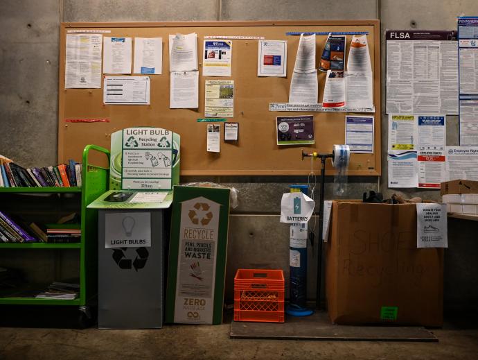 There is a recycling station box with a sign on it that says label "Light bulbs", and next to it there is another container with a sign that has a recycling symbol says "pens markers and pencil waste”. It is standing next to an unmarked milk crate and next to that is a tall cylindrical container for "batteries. There is a large cardboard box next to this which has a sign on it that says “Donate your gently used shoes here”.