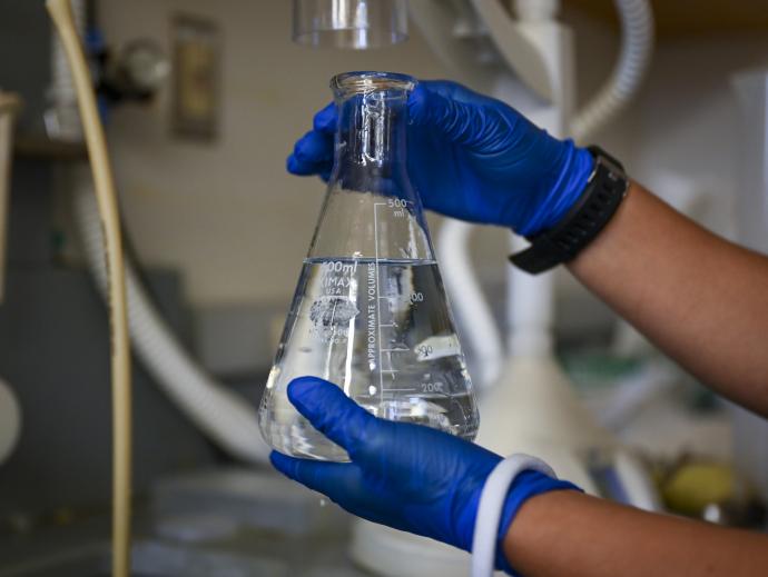 a pair of hands wearing lab gloves are shown holding up a beaker filled with clear liquid in a laboratory setting