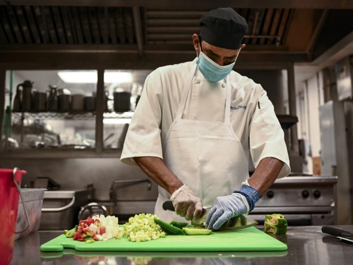 A chef in a professional kitchen is shown wearing a mask while chopping various vegetables on a cutting board with gloved hands.