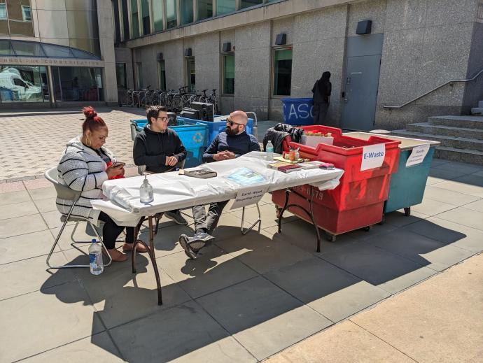 Three people sit at a folding table in a courtyard. There is a sign hanging on the table that reads "Earth Week Recycling Event". Beside the table are two large bins - one is for E-Waste and the other is labeled for "Books, Notebooks, Office Supplies"