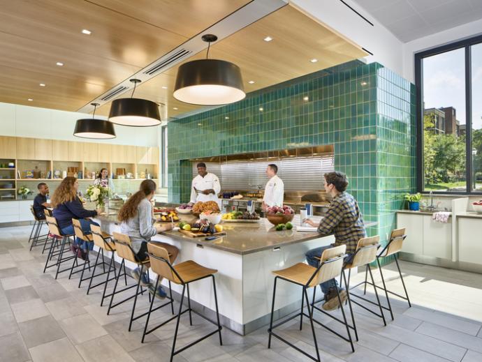 Quaker kitchen at Gutmann college house is shown. It has a large kitchen island with 10 stools around it. On the island, there are several trays of food. Several students are seated around the table. Two people wearing chef uniforms are standing behind the island at the stoves and giving a cooking demonstration.