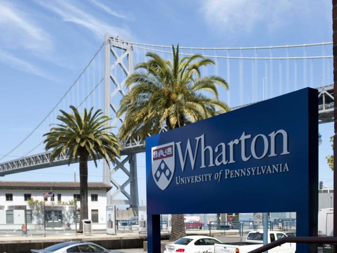 The Wharton Campus sign in the foreground while a bridge and palm trees are behind it